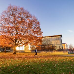 An oak tree in beautiful orange foliage stands before the Skillman Library. A student strolls by.