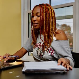 A student sits at a table in Portlock Black Cultural Center accessing a computer and writing in a notebook.