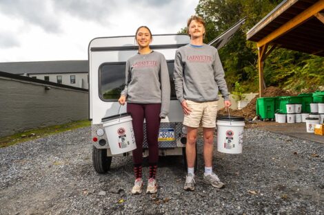 Two students stand in front of an electric vehicle with buckets of compost.