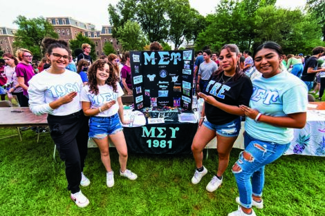 Four students stand in front of their table for their greek organization at a fair.
