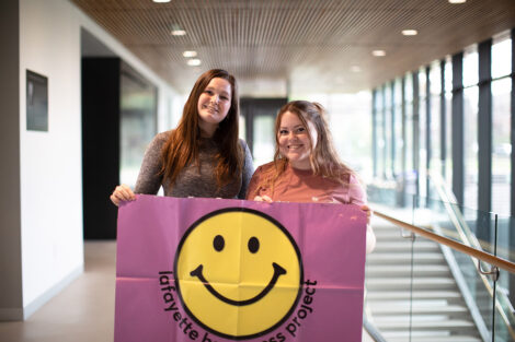 Two students smile holding a flag with a smiley face that reads 