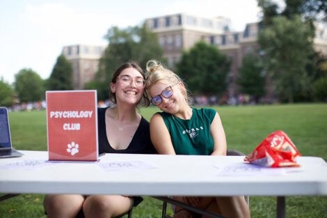 Two students smile at a club fair, with a sign that reads 